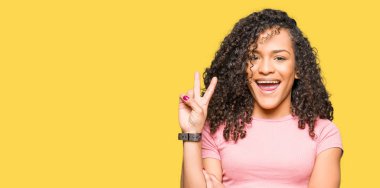 Pembe tişört giyen kıvırcık saçlı Genç güzel kadın mutlu yüz zafer işareti yapıyor kamera göz kırpıyor ile gülümsüyor. İki numara.