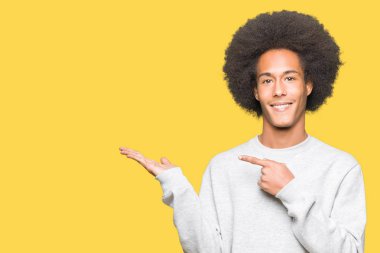 Afro saç ile genç Afrikalı Amerikalı adam sportif Sweatshirt hayran ve kamera için gülümseyen el ile sunmak ve parmak ile işaret ederken.