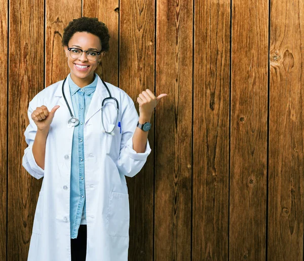 年轻的非洲医生妇女穿着医疗外套在孤立的背景指向后面的背后 手和竖起大拇指 面带微笑自信 — 图库照片