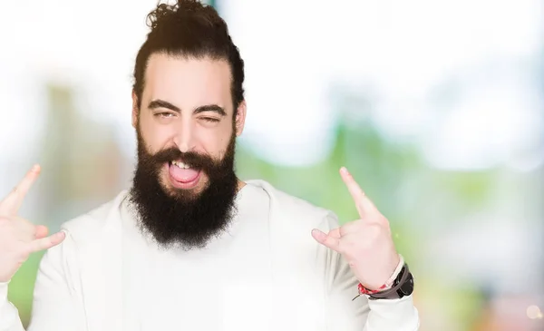 Young man with long hair and beard wearing sporty sweatshirt shouting with crazy expression doing rock symbol with hands up. Music star. Heavy concept.