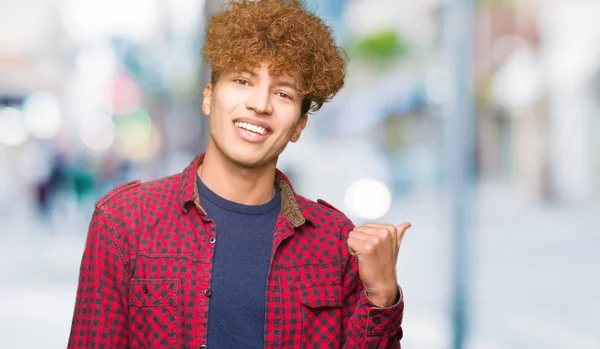 Young handsome student man with afro hair wearing a jacket smiling with happy face looking and pointing to the side with thumb up.