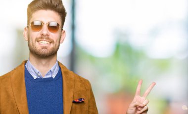 Genç yakışıklı zarif adam mutlu yüz zafer işareti yaparken kameraya göz kırpıyor ile gülümseyen güneş gözlüğü takıyor. İki numaralı.