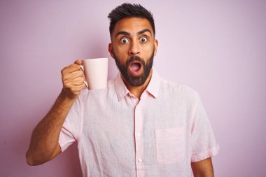 Genç Hintli bir adam gömleğini giyer ve kahve içer. Pembe arka planda durur. Şaşırmış bir yüz ifadesinden korkar ve heyecanlanır.