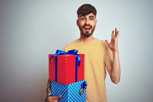 Young man with tattoo holding birthday gifts standing over isolated white background very happy and excited, winner expression celebrating victory screaming with big smile and raised hands