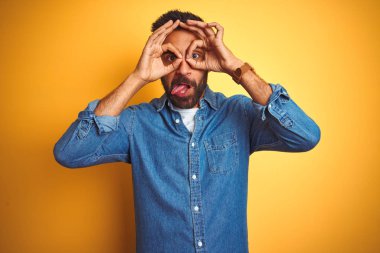 İzole edilmiş sarı arka plan üzerinde duran kot gömlekli genç Hintli adam dürbün gibi jest yapıyor, dili dışarı çıkarıyor, gözleri parmaklarından bakıyor. Çılgın ifade.