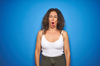 Orta yaşlı, kıvırcık saçlı, mavi yalıtılmış arka planda duran, şaşkın, şüpheci ve alaycı bakışlı, ağzı açık, şaşırmış, orta yaşlı bir kadın.