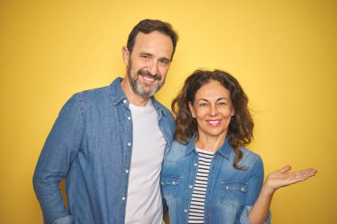 Güzel orta yaşlı çift, izole edilmiş sarı arka planda kot gömlek giyip mutlu bir şekilde sunum yapıyor ve avuç içi kameraya bakıyor..