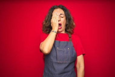 Kırmızı izole arka planda önlük giyen orta yaşlı bir kadın. Yarı yüz, göz ve ağızla esnemekten yorulmuş. Yüzüm acıyor..