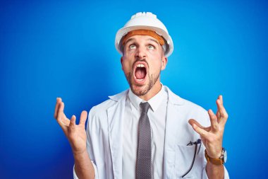 Mavi arka plan üzerinde güvenlik kaskı takan genç, yakışıklı mühendis sinirli ve sinirli bir şekilde bağırıyor ve agresif bir ifadeyle kollarını kaldırıyor. Hayal kırıklığı konsepti.