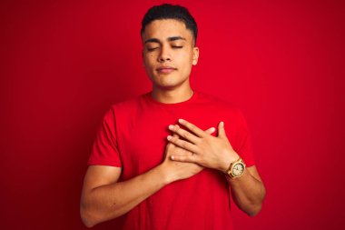 İzole edilmiş kırmızı arka plan üzerinde dikilmiş tişört giyen genç Brezilyalı adam elleri göğsünde, gözleri kapalı ve yüzünde minnet dolu bir jest ile gülümsüyor. Sağlık kavramı.