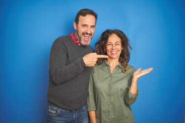 Güzel orta yaşlı çift, izole edilmiş mavi arka planda durarak kameraya hayret ediyor ve gülümsüyor. Elleriyle ve parmaklarıyla işaret ediyor..