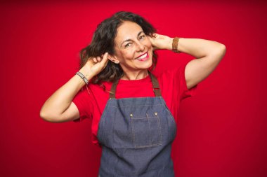 Kırmızı izole edilmiş arka planda önlük giyen orta yaşlı bir kadın parmaklarıyla kulaklarını çekerek gülüyor, komik bir jest yapıyor. Seçme sorunu