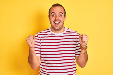 Sıradan çizgili tişört giyen genç adam, izole edilmiş sarı arka planda durup kollarını kaldırarak ve açık gözlerle başarıyı kutluyor ve şaşırıyor. Kazanan konsept.