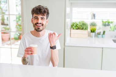 Sabahları kağıt bardakta kahve içen genç bir adam işaret ediyor ve gülümseyen bir yüzle baş parmağıyla onu gösteriyor.