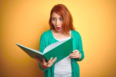 Sarı arka plan üzerine yeşil kitap okuyan genç kızıl saçlı kadın şaşkınlık dolu bir yüzle korkmuş ve korku ifadesinden heyecanlanmış.