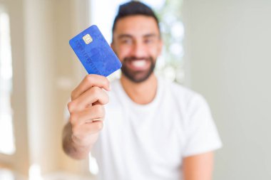 İspanyol yakışıklı duran ve diş gösterilen kendine güvenen bir gülümsemeyle gülümseyerek mutlu bir yüz ile kredi kartı tutan