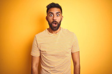 İzole edilmiş sarı arka plan üzerinde duran genç Hintli adam şaşkınlık ve şaşkınlık dolu ifade, korku ve heyecan dolu yüz ifadesiyle dehşete düşmüş ve şok olmuştu..