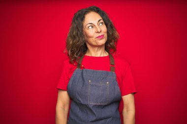 Kırmızı izole edilmiş arka planda önlük giymiş orta yaşlı bir kadın gülümsüyor ve başka yöne bakıyor..