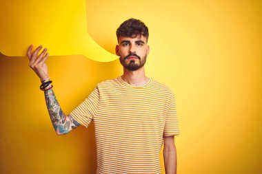 Dövmeli genç bir adam konuşma baloncuğu tutarak izole edilmiş sarı arka planda duruyor. Yüzünde kendine güvenen bir ifade var.