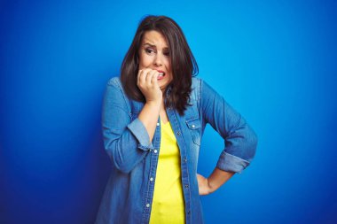 Genç güzel esmer kadın mavi izole arka plan üzerinde rahat mavi denim ceket giyen vurguladı ve ağız ısırma çivi eller ile gergin bakıyor. Anksiyete sorunu.