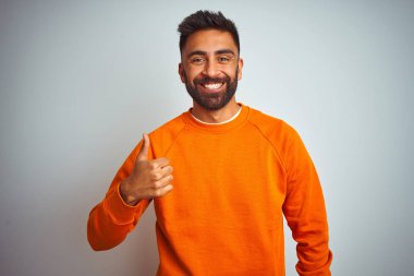İzole edilmiş beyaz arka planda turuncu süveter giyen genç Hintli adam mutlu bir el hareketi yapıyor. Kameraya bakıp başarıyı gösteren ifade onaylanıyor.