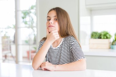 Çizgili tişört giyen güzel genç kız çocuk, çapraz kolları ve çenesi üzerinde kaldırılan elleri ile gülümseyerek kameraya güvenle bakıyor. Olumlu düşünme.