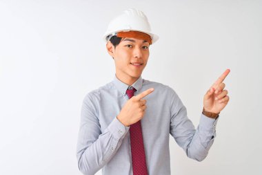 Kravat ve miğfer takan Çinli mimar izole edilmiş beyaz arka planda duruyor gülümsüyor ve kameraya bakıyor iki eliyle ve parmaklarıyla yan tarafı işaret ediyor..