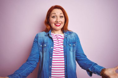 Güzel kızıl saçlı kadın kot gömlek ve çizgili tişört giyip pembe arka planda mutlu bir şekilde gülümseyerek dostça hoş geldin, pozitif ve kendinden emin selamlar.