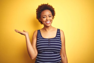Beauitul afro-amerikan kadın izole sarı arka plan üzerinde yaz t-shirt giyen neşeli sunan gülümseyerek ve kameraya bakarak avuç içi ile işaret.