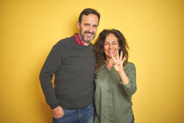 Güzel orta yaşlı çift, izole edilmiş sarı arka planda dört numaralı parmaklarıyla gülümserken ve mutlu bir şekilde gülümserken görülüyor..