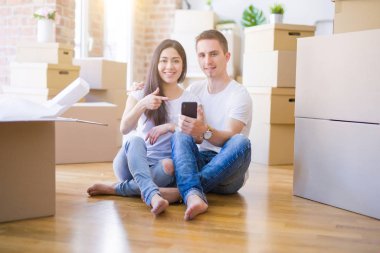 Karton kutuların etrafında akıllı telefonlar kullanarak yerde oturan güzel bir çift mutlu bir şekilde elleri ve parmaklarıyla işaret ediyorlar.