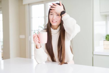 Bir fincan kahve içen güzel Asyalı kadın, elleri başının üstünde, utancından şoke olmuş ve şaşırmış, sinirli ve hayal kırıklığına uğramış. Hata için korku ve üzüntü.