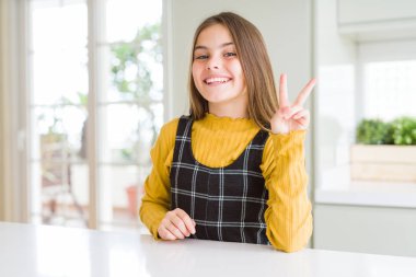 Genç, güzel sarışın kız evde sarı süveter giyiyor ve iki numaralı parmaklarıyla onu gösterirken kendinden emin ve mutlu bir şekilde gülümsüyor..