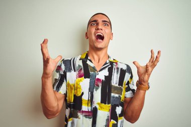 Beyaz arka plan üzerine yaz tişörtü giyen yakışıklı genç adam kızgın ve çılgın bir şekilde bağırıyor ve agresif bir ifade ve kollarla bağırıyor. Hayal kırıklığı konsepti.