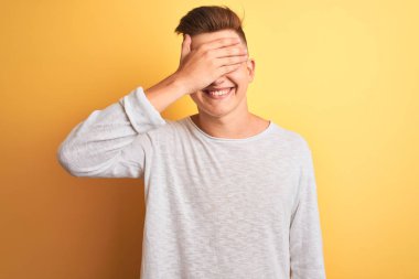 İzole edilmiş sarı arka plan üzerinde duran beyaz tişörtlü yakışıklı bir adam. Gülümsüyor ve elleri yüzünde şaşkınlıktan gözleri kapatıyordu. Kör kavram.