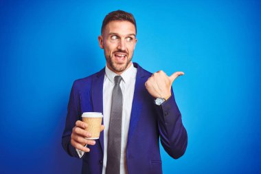 Genç yakışıklı iş adamı, mavi izole bir arka planda kağıt bardakta kahve içiyor ve gülümseyen bir gülümsemeyle baş parmağıyla onu işaret ediyor.