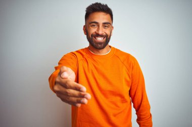 İzole edilmiş beyaz arka planda turuncu süveter giyen genç Hintli adam selamlaşma ve hoş geldin demek için gülümseyerek tokalaşıyor. Başarılı bir iş..
