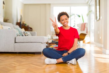 Genç ve güzel Afro-Amerikalı kadın evinde oturmuş kameraya bakıp zafer işareti yaparken gülümsüyor. İki numara..