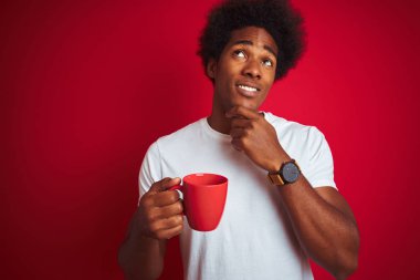 Genç Afrikalı Amerikalı bir adam bir fincan kahve içerken izole edilmiş kırmızı arka planda dikiliyor ciddi bir yüz sorgulama düşünüyor, kafası çok karışık bir fikir.