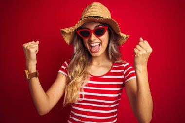 Kırmızı izole arka planda güneş gözlüğü ve yaz şapkası takan genç ve güzel bir kadın kollarını kaldırarak ve açık gözlerle başarıyı kutluyor ve şaşırıyor. Kazanan konsept.
