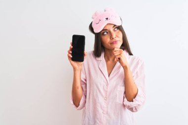 Uyku maskesi takan genç bir kadın, elinde akıllı telefon, izole edilmiş beyaz arka planda ciddi bir yüz, soru soran, kafası karışık bir fikir.