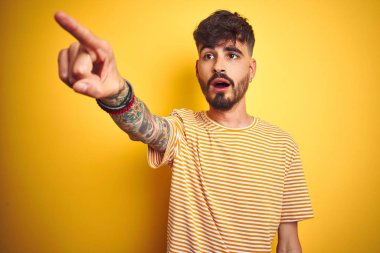 Dövmeli, çizgili tişörtlü genç adam izole edilmiş sarı arka planda dikiliyor. Parmağı önde, ağzı açık ve şaşırmış bir şekilde işaret ediyor.