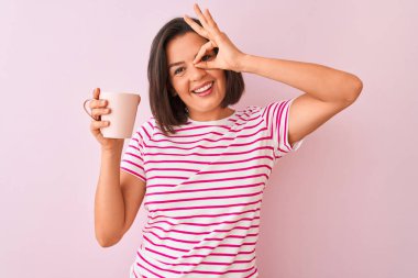 Genç ve güzel bir kadın izole edilmiş pembe arka planda durup kahve içiyor. Gülümsüyor ve el pençe divan duruyor.