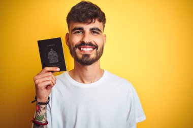 Kanada pasaportu takan genç bir adam izole edilmiş sarı arka planda mutlu bir yüzü olan ve kendine güvenen bir gülümsemeyle dişlerini gösteren.