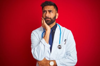 Steteskop takan genç Hintli doktor, izole edilmiş kırmızı arka planda duruyor. Kollarını çapraz tutmaktan sıkılmış ve yorgun görünüyor..