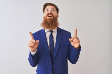 İzole edilmiş beyaz arka plan üzerinde duran genç, kızıl saçlı iş adamı şaşırmış ve şaşırmış bir şekilde yukarı bakıyor ve parmaklarıyla işaret ediyor ve kollarını kaldırıyordu..