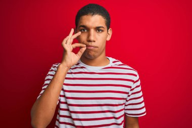 İzole edilmiş kırmızı arka plan ağzının üzerine çizgili tişört giyen yakışıklı Arap adam ve dudakları parmaklarıyla kapanan fermuar gibi. Gizli ve sessiz, tabu konuşması