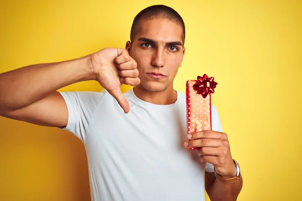 黄色の隔離された背景の上に誕生日プレゼントを持っている若い白人男性怒りの顔を持ちます 親指で嫌いを示す負の記号ダウン 拒絶概念 — ストック写真