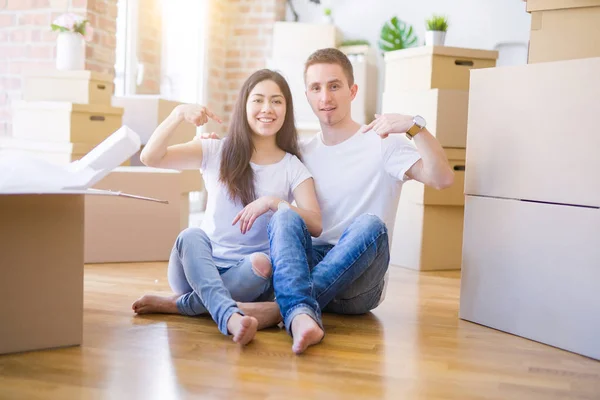 Young beautiful couple sitting on the floor at new home around cardboard boxes with surprise face pointing finger to himself
