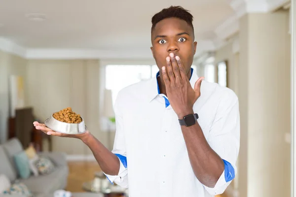 African american man holding metal bowl with cat or dog dry food cover mouth with hand shocked with shame for mistake, expression of fear, scared in silence, secret concept